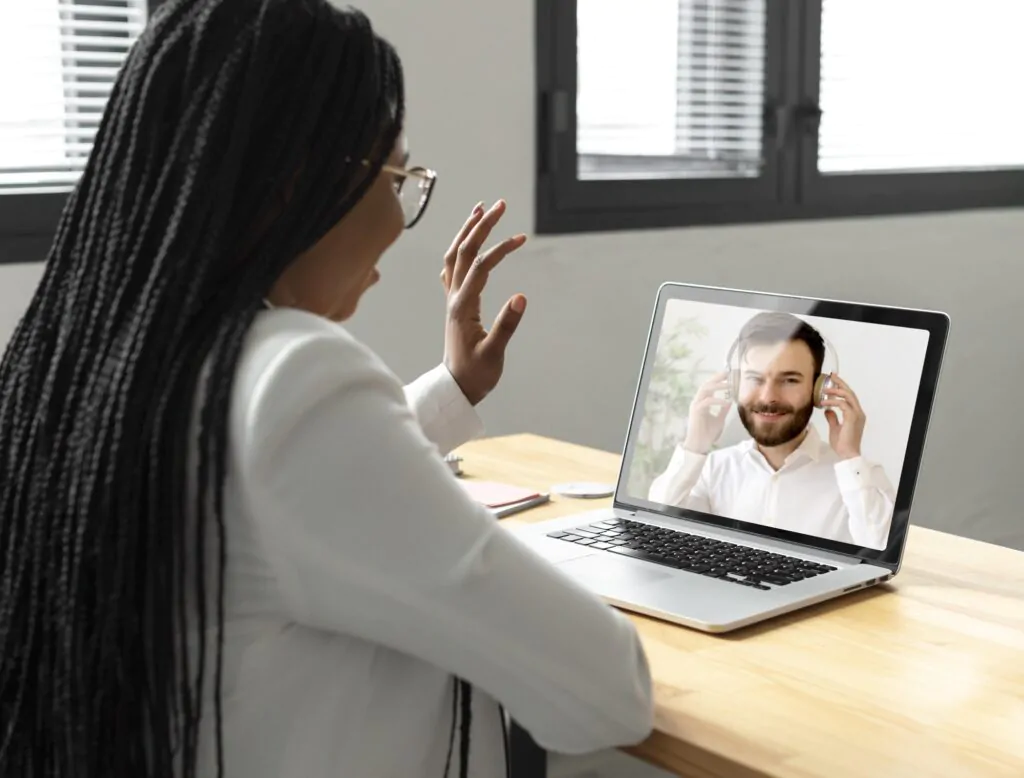 Na imagem, uma mulher com cabelos longos trançados e óculos está sentada à mesa em frente a um notebook, participando de uma videoconferência. Ela usa uma roupa branca formal e está gesticulando com a mão direita enquanto fala. Na tela do notebook, aparece um homem sorridente com barba curta e fones de ouvido brancos, vestindo uma camisa branca. Ele parece ouvir atentamente e ajustar os fones de ouvido com ambas as mãos. A mesa é de madeira clara e possui alguns objetos, como um celular e um caderno. O ambiente é iluminado com luz natural vinda das janelas com persianas parcialmente fechadas ao fundo, criando um ambiente profissional e neutro.